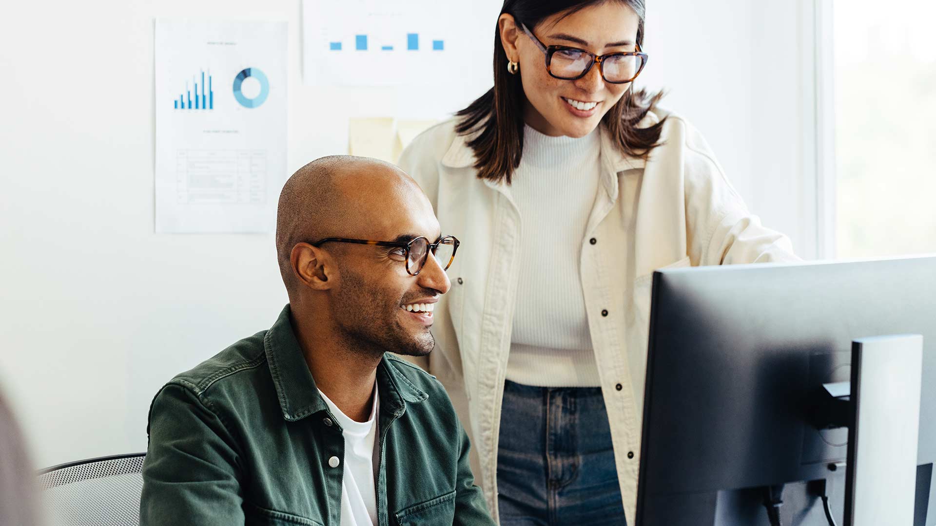 A man and woman smiling while looking at a computer screen, suggesting collaboration. A graph and chart on the wall hint at a professional setting.